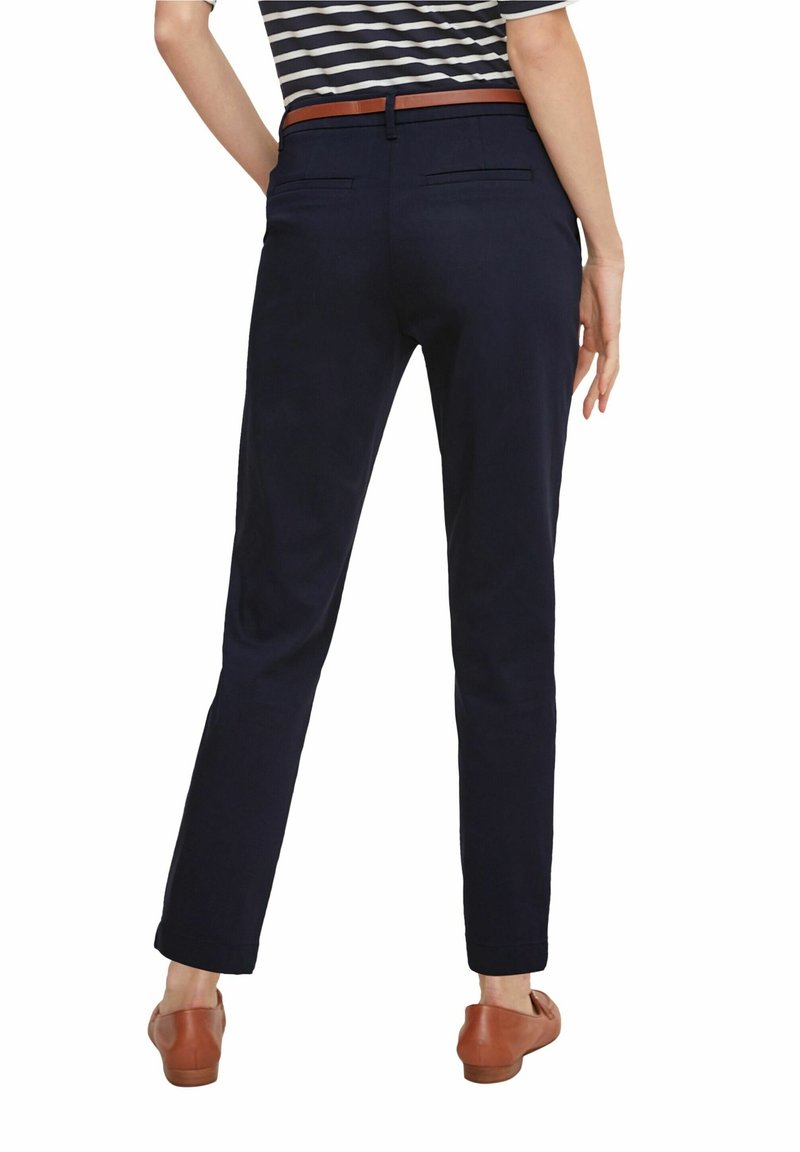 Femme portant un pantalon slim bleu marine foncé, une ceinture marron et des chaussures plates marron, debout de dos face au spectateur sur un fond blanc.