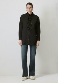 Zwarte overhemd met drie grote zwarte strikaccenten aan de voorkant, lange mouwen en een klassieke kraag, gecombineerd met uitlopende blauwe jeans.
