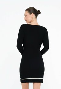 Donna con capelli scuri raccolti in un chignon, che indossa un abito aderente a maniche lunghe di colore nero con un bordo bianco vicino all'orlo, girata di spalle su uno sfondo neutro.