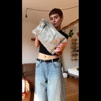 Imballaggio in plastica trasparente che contiene un oggetto marrone. La persona indossa una maglietta nera e jeans blu larghi, in piedi in uno spazio interno ben illuminato.
