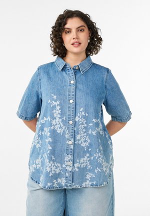 Kvinde med krøllet hår iført en blå kortærmet denimskjorte med hvide blomsterprint og lyse denimbukser.