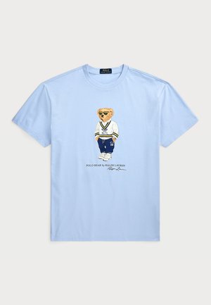 Camiseta de algodón azul claro con un gráfico de un oso vestido con ropa deportiva, con el texto "POLO BEAR by RALPH LAUREN" debajo.