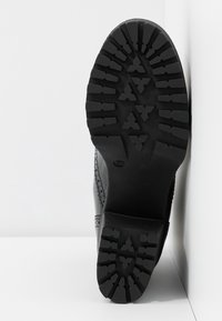 Chaussure en cuir noire avec une semelle en caoutchouc texturée. La semelle présente un motif de crampons robuste pour une meilleure adhérence. Le côté est orné de perforations décoratives.