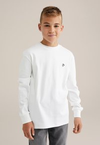 Vit långärmad t-shirt i bomull, med rund halsringning och ett litet svart handemblem på vänster bröst. I kombination med grå jeans.