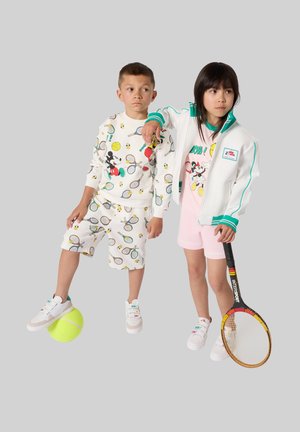 Two children in tennis-themed clothing pose; one balances a large tennis ball on foot, the other holds a tennis racket.