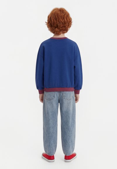 Dziecko z kręconymi rudymi włosami stoi tyłem, ubrane w niebieską bluzę z czerwonymi wykończeniami, jasnoniebieskie dżinsy i czerwone trampki.