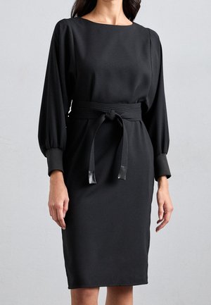 Frau trägt ein langärmeliges schwarzes Kleid mit rundem Ausschnitt und einem breiten Gürtel, der in der Taille zu einem Knoten gebunden ist.