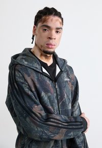 Camo zip-up hoodie with black and green leaf patterns, featuring gray accents and three black stripes on sleeves. Soft fabric texture.