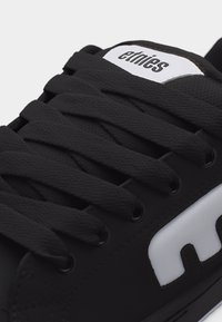 Μαύρο αθλητικό παπούτσι με μαύρα κορδόνια, λευκό λογότυπο «etnies» στη γλώσσα και λευκή λεπτομέρεια στο πλάι σε σχήμα γράμματος «E».