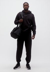 Homme portant un sweat à capuche noir, un pantalon de survêtement et des baskets, portant un sac de sport noir sur l'épaule, debout devant un fond blanc.
