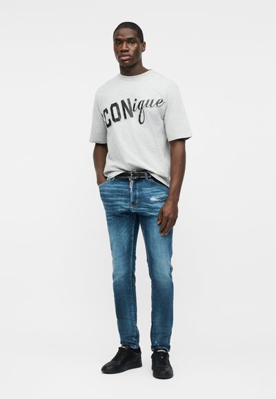 Άνδρας στέκεται με τα χέρια στις τσέπες, φορώντας γκρι μπλούζα με το λογότυπο "ICONique", μπλε τζιν, μαύρη ζώνη και μαύρα αθλητικά παπούτσια, σε απλό φόντο.
