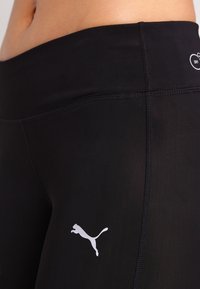 Czarne legginsy sportowe wykonane z elastycznego materiału. Mają wysoki stan i białe logo Pumy po lewej stronie. Gładka textura.