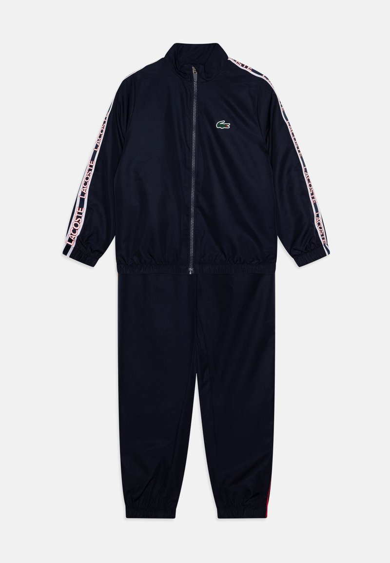 Fato de treino azul-marinho com casaco com fecho e punhos elásticos. Apresenta detalhes laterais em riscas rosadas e brancas e um pequeno logo verde no peito.