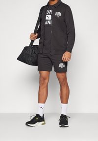 Conjunto atlético negro que incluye una chaqueta con cremallera, camiseta, pantalones cortos y zapatillas. Presenta acentos de logotipo y calcetines blancos en contraste.