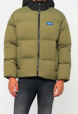 Homme portant une doudoune vert olive avec capuche noire, fermeture éclair frontale et étiquette bleue "HUGO" sur la poitrine, associée à un jean foncé.