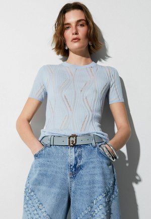 Donna con maglia a maniche corte azzurra chiara con motivi ondulati, jeans in denim azzurro chiaro a vita alta, cintura e braccialetti d'argento, mani in tasca.