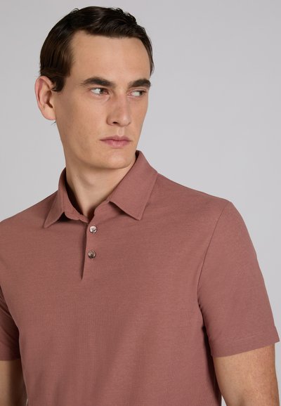 Jeune homme aux cheveux courts et foncés, portant un polo à manches courtes couleur rose poussiére, regardant vers sa gauche sur un fond gris uni.