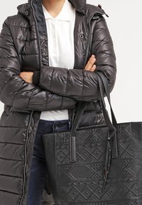 Osoba nosící černou prošívanou bundu přes bílou polo triko, držící velkou černou vzorovanou tašku s překříženýma rukama.