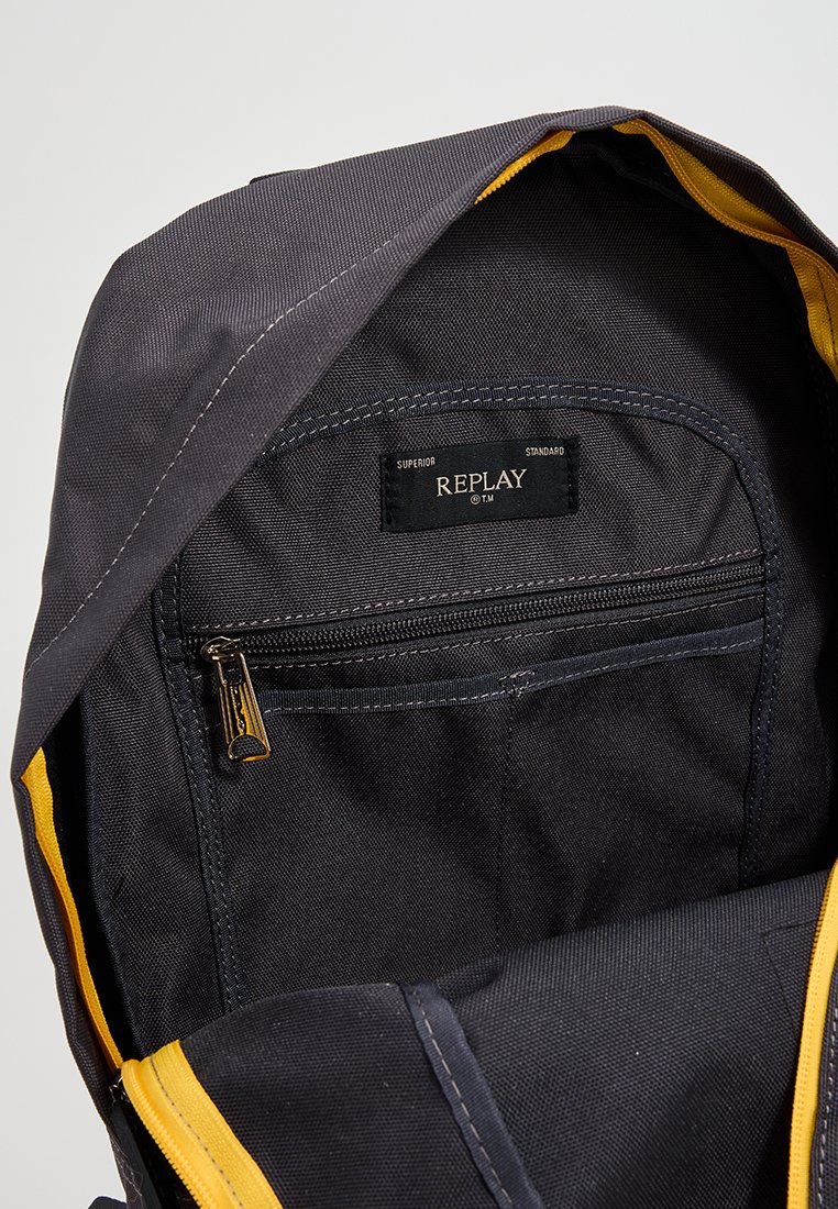 Interno di uno zaino in tessuto nero con una foderatura gialla, una tasca con zip e un'etichetta con la scritta "REPLAY". Texture durevole con design strutturato.