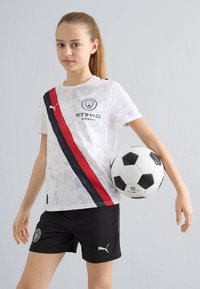 Baltas „Manchester City“ marškinėliai su raudona ir juoda įstrižąja juosta, su logotipu ir užrašu „Etihad Airways“. Juodos kelnaitės su „Puma“ logotipu; laikantis futbolo kamuolį.