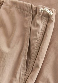 Beige stofbukser med en blød tekstur, udstyret med en snor i taljen og synlig hvid syning langs kanterne og lommerne.