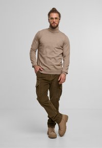 Mann mit Brille, der einen beigen Rollkragenpullover, braune Cargo-Hosen und tanfarbene Stiefel trägt, steht mit einer Hand in der Tasche vor einem schlichten Hintergrund.