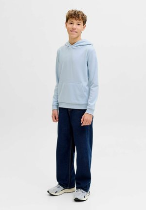 Adolescente de pie, vistiendo una sudadera con capucha azul claro, vaqueros azul oscuro y zapatillas blancas, sobre un fondo blanco liso.