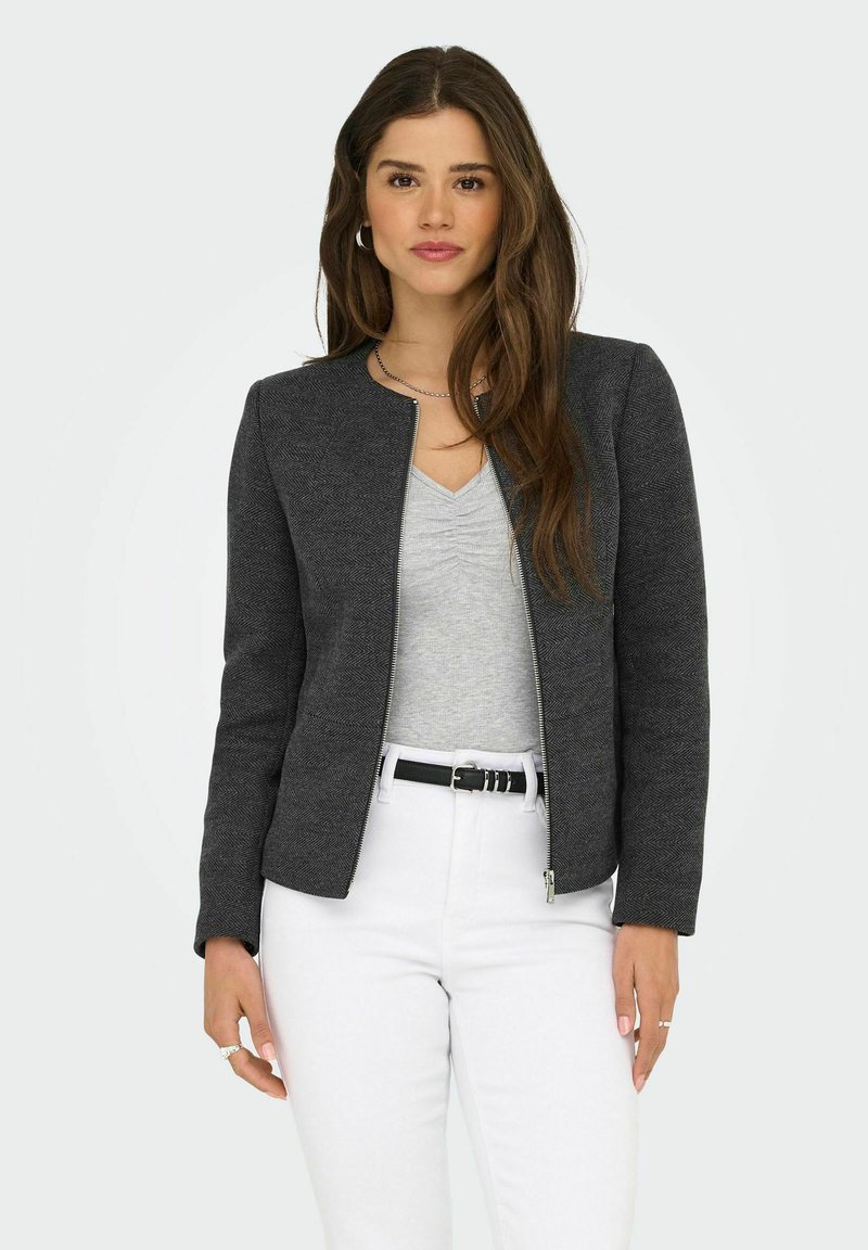 Veste zippée à chevrons gris avec un col rond, associée à un haut gris et un pantalon blanc, arborant une coupe ajustée.