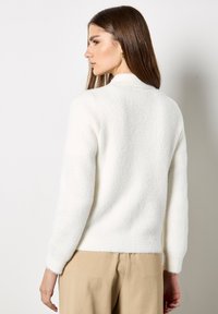 Donna con lunghi capelli castani, che indossa un maglione bianco morbido e pantaloni beige, di profilo contro uno sfondo chiaro neutro.