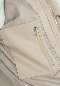 Beige jacka i närbild som visar insidan av fickan med dragkedja och ett slätt foder med synliga stygn och metalldragkedja.