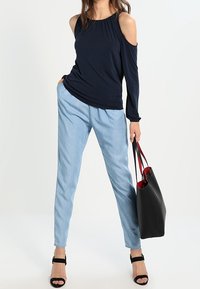 Námořnicky modrý top s odhalenými rameny, světle modré zúžené kalhoty, černá taška přes rameno s červenými akcenty a černé páskové podpatky na neutrálním pozadí.