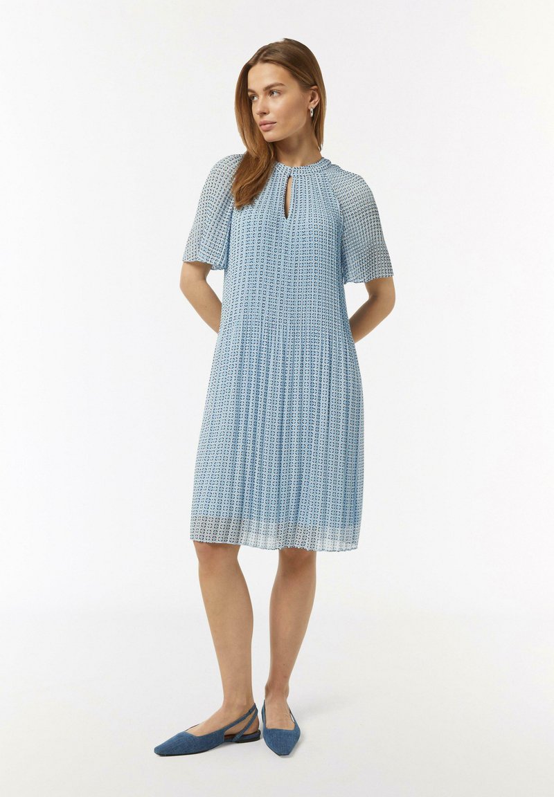 Lichtblauwe jurk met korte mouwen en een ronde halslijn met sleutelgatdetail, voorzien van een patroontextuur en plooien. Draagt met blauwe ballerina's.
