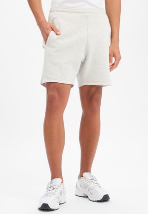 Mann trägt hellgraue Sportshorts und weiße Laufschuhe, steht mit einer Hand in der Tasche vor weißem Hintergrund.