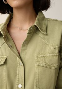 Olivgrünes Denimhemd mit spitzem Kragen, Druckknöpfen und zwei Brusttaschen; zeigt sichtbare Textur- und Nähdetails.