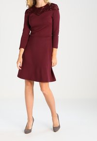 Mujer vistiendo un vestido burdeos a la altura de la rodilla con detalles de encaje en el pecho superior y las mangas, combinado con zapatos de tacón alto grises.