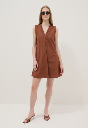 Mujer con vestido marrón sin mangas con escote en V con volantes, sandalias marrones de tacón y gafas de sol negras, de pie frente a un fondo liso y claro.