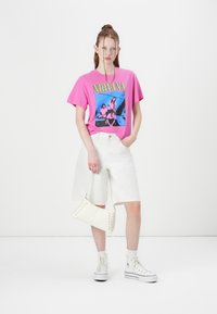 Rosa grafisk t-skjorte med banddesign, hvite denimshorts, hvit crossover-veske med sømdetaljer, og hvite høye sneakers.