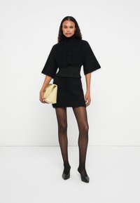 Μαύρος φουντωτός oversized πουλόβερ με φαρδιά μανίκια, συνδυασμένο με εφαρμοστό μαύρο μίνι φόρεμα και διαφανείς μαύρες καλσόν. Κρατάει μια ανοιχτό κίτρινη τσάντα.