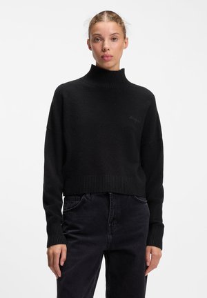 Donna che indossa un maglione nero a coste con collo alto e jeans neri a vita alta, in piedi davanti a uno sfondo bianco uniforme.