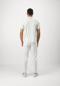 Polo vert menthe à manches courtes, en tissu léger avec un col rond et un logo noir dans le dos, associé à un pantalon blanc et des chaussures de sport.