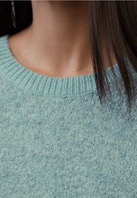 Hellblauer Pullover mit strukturierter Oberfläche, ausgestattet mit einem gerippten Rundhalsausschnitt und weichem Strickstoff. Nahaufnahme des Kragenbereichs und der Haare.