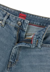 Blå denimjeans med en HUGO-märkesetikett, mässingsknapp och dragkedjestängning. Anmärkningsvärd röd sömnad inuti midjebandet och dragkedjeområdet.