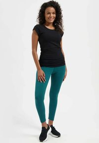 Frau mit lockigem Haar, die ein schwarzes Kurzarmshirt, türkisfarbene Leggings und schwarze Turnschuhe trägt, steht lächelnd vor einem weißen Hintergrund.