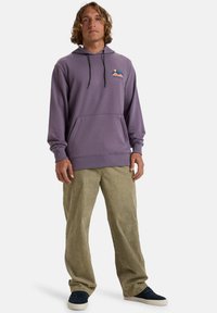 Purple hoodie with a front pocket, adjustable drawstring hood, and graphic of mountains. Paired with light green corduroy pants and navy shoes.