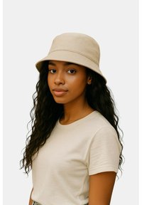 Beige stoffen emmerhoed met brede rand en zachte textuur, gedragen met een lichtgekleurde T-shirt. Platte achtergrond.