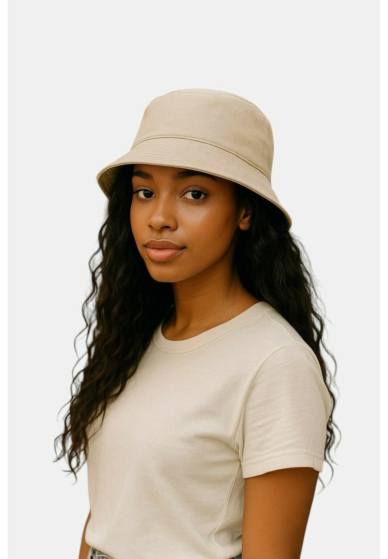 Beige stoffen emmerhoed met brede rand en zachte textuur, gedragen met een lichtgekleurde T-shirt. Platte achtergrond.