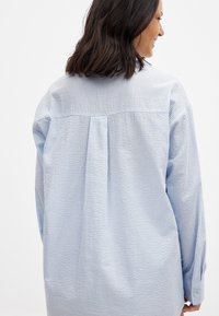 Blue and white striped button-up shirt with a back pleat, long sleeves, and a straight hem. Cotton fabric with a textured finish.