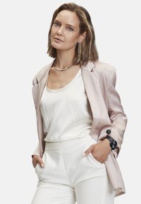 Hellrosa Satin-Blazer über einem weißen Oberteil, kombiniert mit weißen Hosen. Silberne Halskette und Ohrringe, schwarzes Armband mit Mesh-Band. Strukturiertem Stoffdetails.