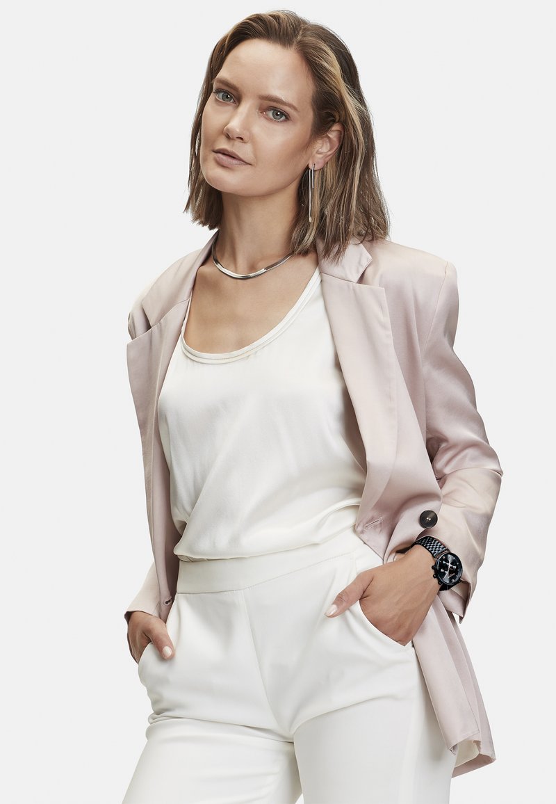 Hellrosa Satin-Blazer über einem weißen Oberteil, kombiniert mit weißen Hosen. Silberne Halskette und Ohrringe, schwarzes Armband mit Mesh-Band. Strukturiertem Stoffdetails.