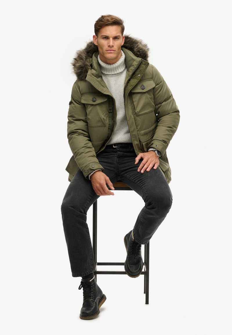 Chaqueta de plumas verde oliva con capucha de pelo sintético, dos bolsillos frontales, llevada sobre un suéter gris claro y combinada con jeans negros y botas.